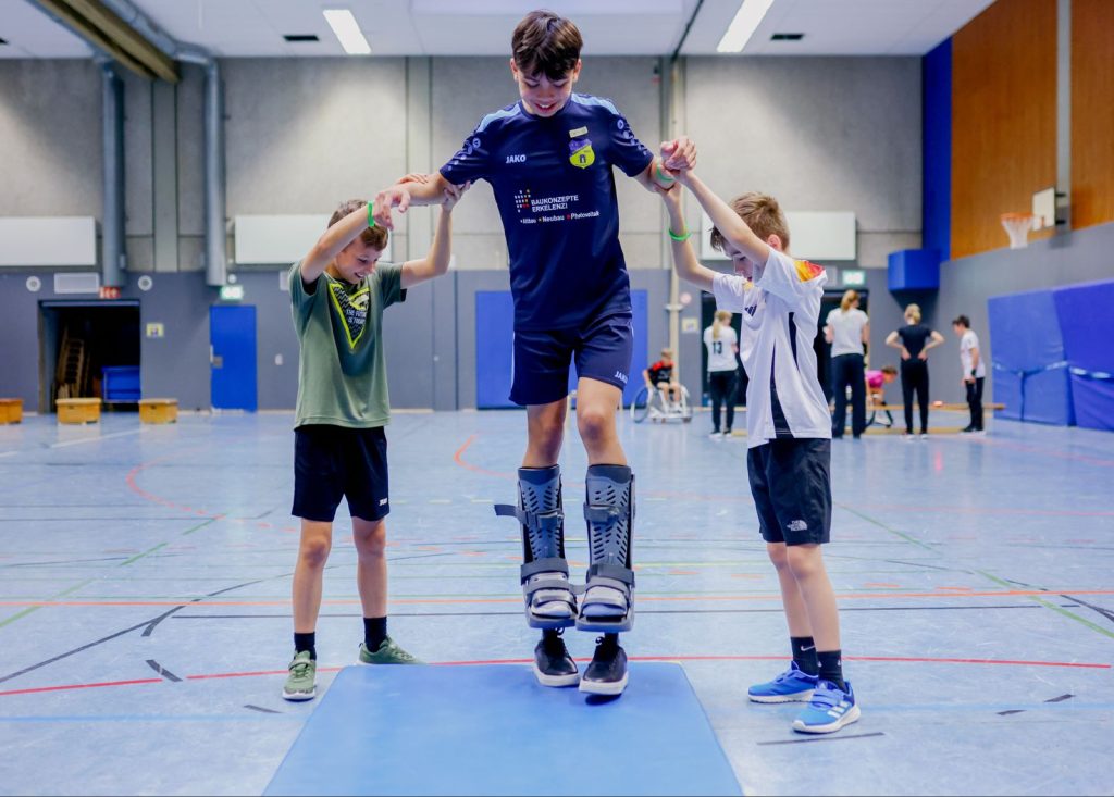 Ein Junge versucht das gehen auf Sportprothesen, zwei weitere Jungs stützen ihn links und rechts.